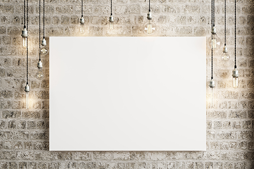 Photo of a blank canvas hanging on a brick wall with exposed lightbulbs around it, suspended from the ceiling.