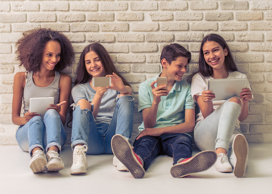 Photo of teenagers with gadgets sitting on the floor against a brick wall.