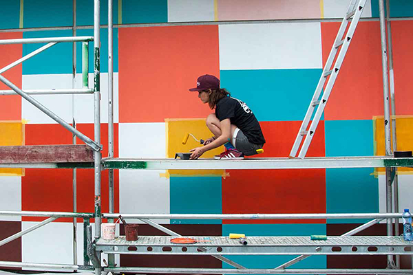 Woman on a scaffolding, painting the exterior of a prison with bright colors.