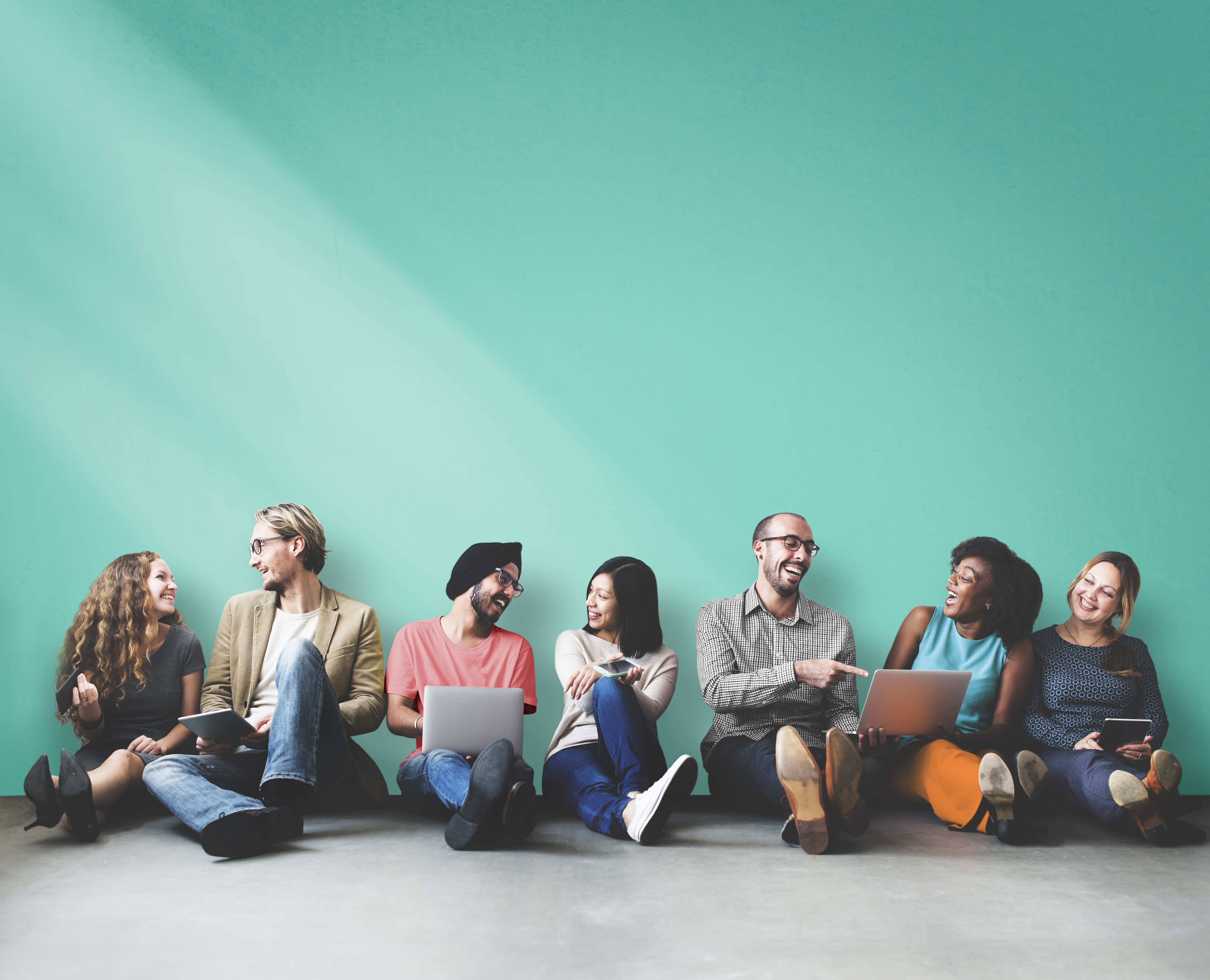 A diverse group of individuals sitting against a blue wall, engaged in conversation and using laptops