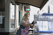 Photo of lady wearing a mask while wheeling a large cart of food from a store to the street.
